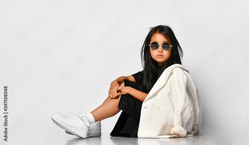 Naklejka premium Side view of little stylish girl model posing in studio wearing sunglasses and looking straight. The girl is dressed in a black dress, white jacket and sneakers. Stylish clothes for schoolchildren.