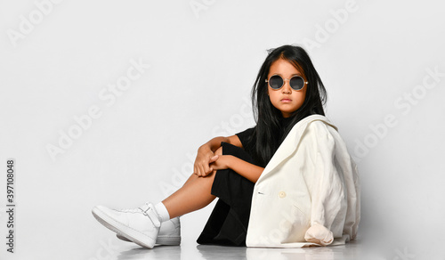 Side view of little stylish girl model posing in studio wearing sunglasses and looking straight. The girl is dressed in a black dress, white jacket and sneakers. Stylish clothes for schoolchildren.