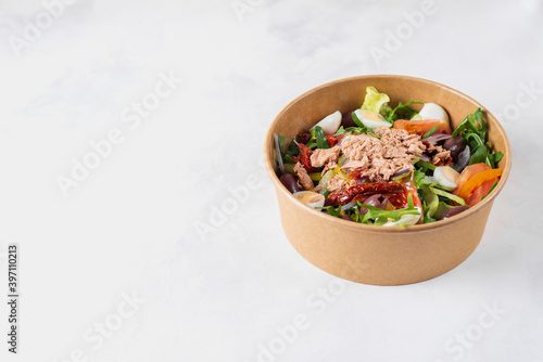 Fish diet salad with tuna, tomatoes, arugula on light background top view. Healthy lunch. Eco-friendly carton packaging, environment protection. Food delivery in disposable plate of craft paper.