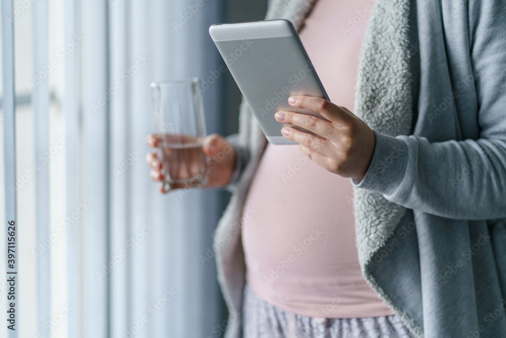 Close u on hand of unknown caucasian woman pregnant mother holding glass of water and digital tablet while standing by the window - pregnancy and water drinking concept Close u on hand of unknown caucasian woman pregnant mother holding glass of water and digital tablet while standing by the window - pregnancy and water drinking concept