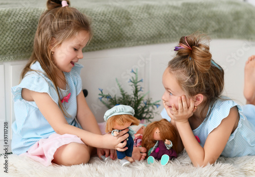 Cheerful pretty small girls sisters kids with long hair in same clorhing playing with toys dolls together having fun at home. Happy childhood, cheerful lifestyle, games, comfortable pastime at home photo