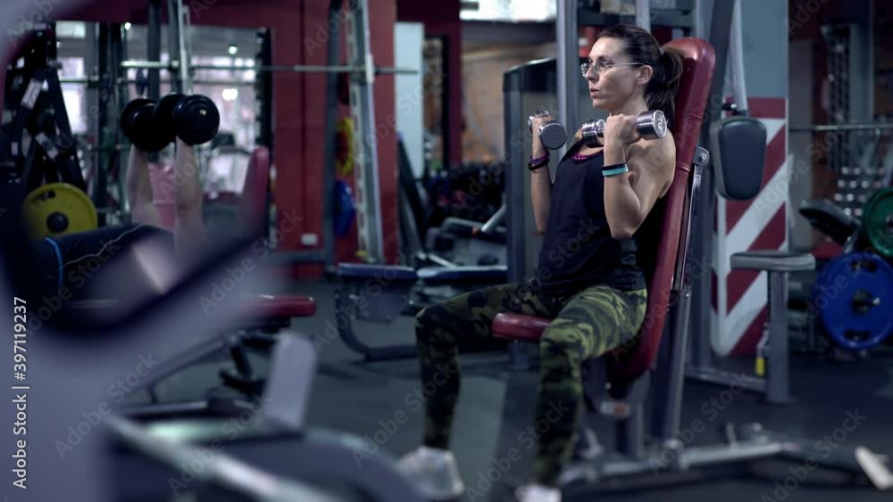 30s or 40s lady in good shape sitting, holding dumbbells in arms and energetically pumping muscles. Portrait of middle-aged female doing physical exercise with dumbbells in gym. Health concept. 30s or 40s lady in good shape sitting, holding dumbbells in arms and energetically pumping muscles. Portrait of middle-aged female doing physical exercise with dumbbells in gym. Health concept.