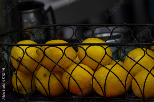 A black wire basket filled with large whole bright yellow organic lemons sits on a table in a restaurant kitchen. The black metal container has tasty lemon citrus fresh fruit from a recent harvest.