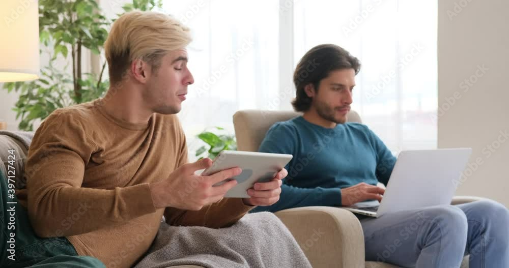 Gay man sharing media content on digital tablet with boyfriend who is using laptop at home Gay man sharing media content on digital tablet with boyfriend who is using laptop at home