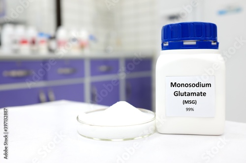 Selective focus of bottle of pure monosodium glutamate or msg food additive beside a petri dish with white solid powder substance. White laboratory background with copy space. photo