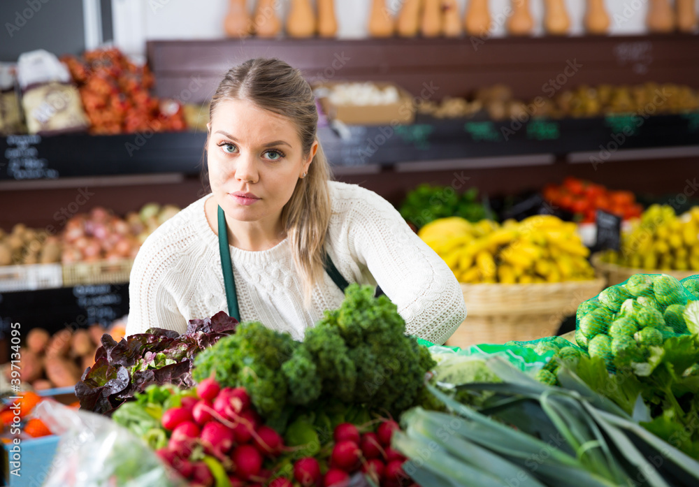 Young female grocery worker arranging fresh greens on store shelves
