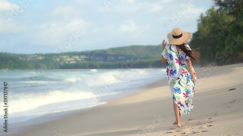 A woman in a colorful print coverup and straw sun hat walks along a windswept beach as big waves crash along the shore. Copy space photo