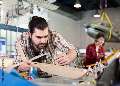 Young diligent cheerful handsome bearded man enjoying his hobby - modeling light airplanes in aircraft hangar