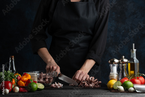 Chef cuts with knife boiled octopuses for cooking traditional asian dish. Backstage of preparing seafood on dark blue background. Concept of exotic asian cuisine. Frozen motion.