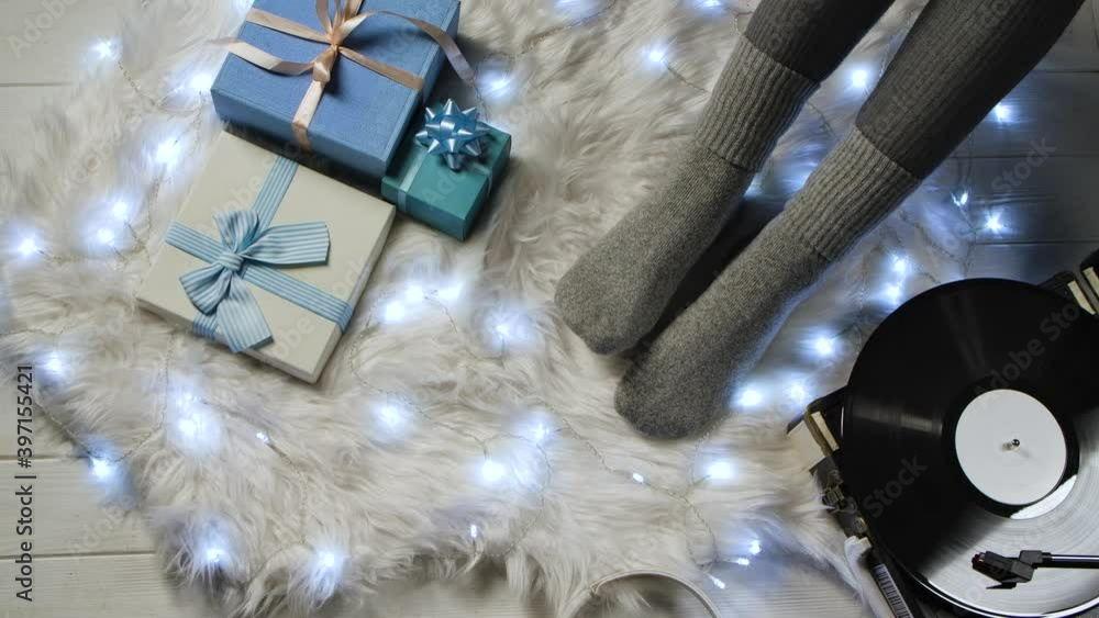 Female feet in gray socks next to blue gift boxes, headphones, luminous garland and retro turntable vinyl. Woman relaxes on white fur. Home comfort concept. Close up. Slow motion.
