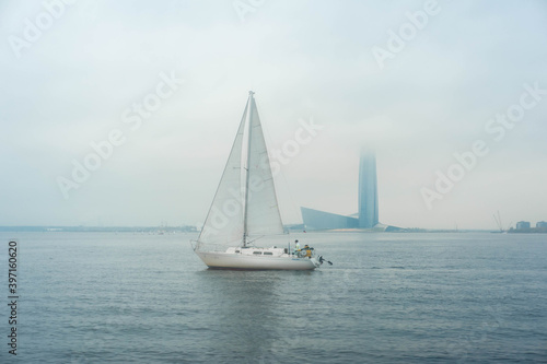  Sailboat in the waters of the Gulf of Finland. Saint-Petersburg, Russia. Foggy cloudy day, blue sky with low clouds and blue water. © Наталья Канищева