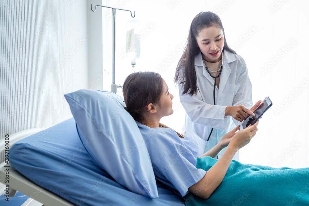Asian female doctor therapeutic advising with tablet to young female patient on Bed in hospital background.