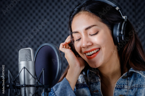 Happy cheerful pretty smiling of portrait a young Asian woman vocalist Wearing Headphones recording a song front of microphone in a professional studio