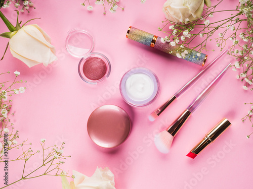 Shiny make up products and accessories on pink background with flowers. Trendy holographic brushes.