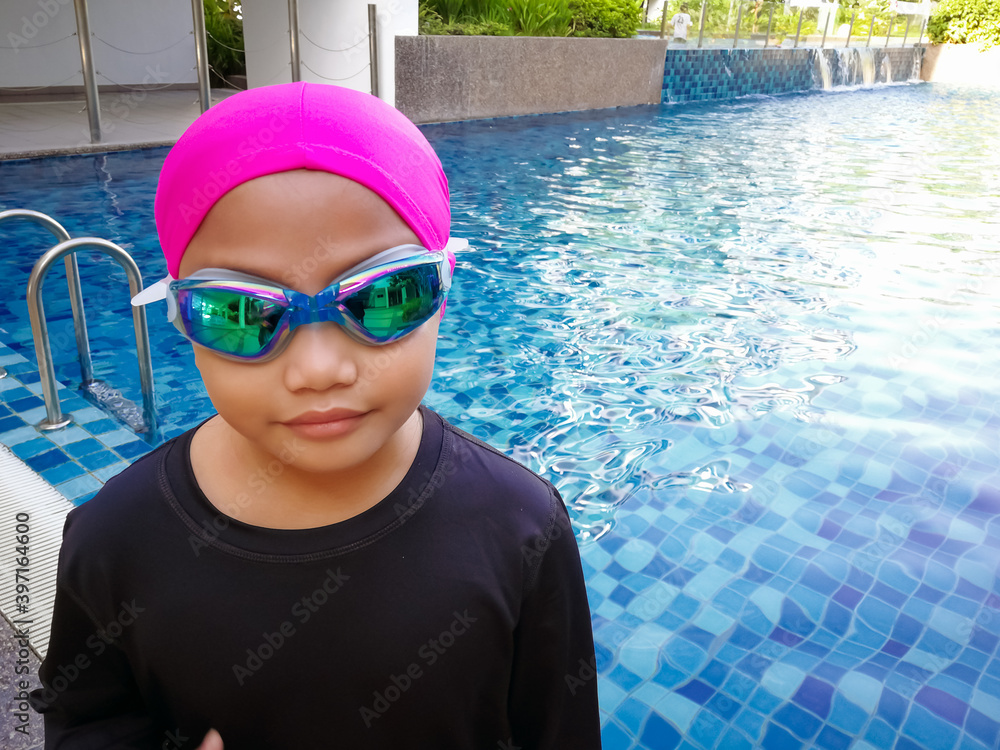 Portrait of a young Asian girl in goggles and swimming cap with swimming pool in the background.