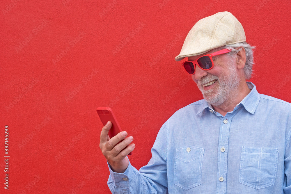 Portrait of senior white haired man with cap smiling using his smart phone, standing against red background Portrait of senior white haired man with cap smiling using his smart phone, standing against red background