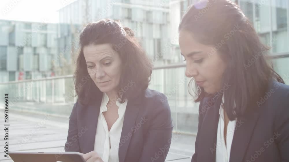 Serious businesswomen using laptops for work together while sitting near office building outside, discussing content and pointing at displays. Business communication and teamwork concept. Serious businesswomen using laptops for work together while sitting near office building outside, discussing content and pointing at displays. Business communication and teamwork concept.