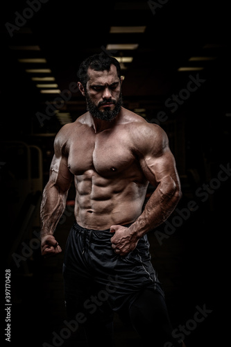 strong young bearded athlete male with perfect sport physique body torso muscle in dark fitness gym