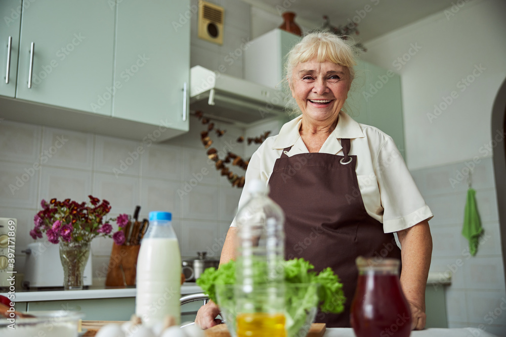 Joyful elderly woman in apron standing in kitchen Joyful elderly woman in apron standing in kitchen
