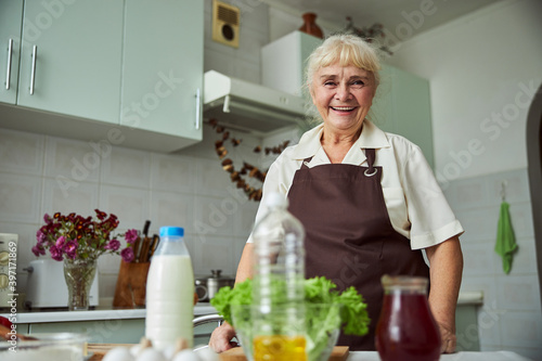 Joyful elderly woman in apron standing in kitchen