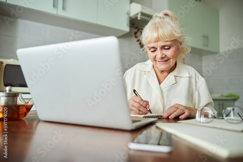 Joyful old woman using modern notebook and taking notes