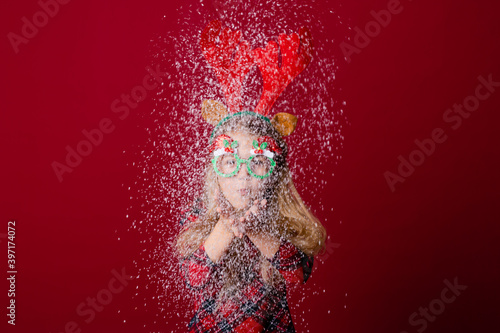 Fototapeta Naklejka Na Ścianę i Meble -  cute little girl in Christmas glasses blows snow from palms in the studio on a red background. Christmas concept, text space