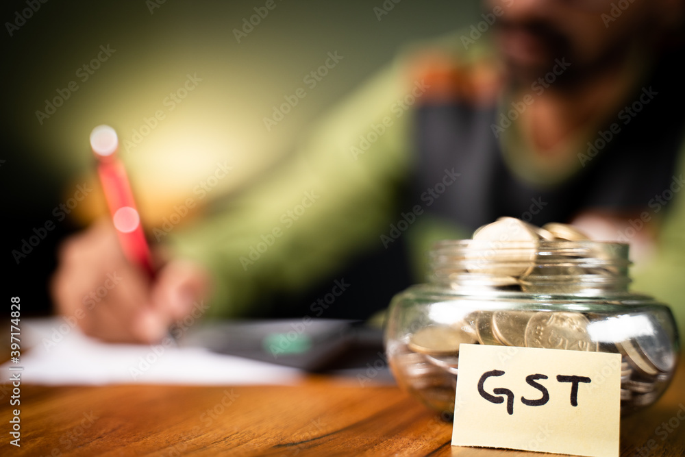 selective focus on coins in bottle and GST sticky note, man in background checking spending's on calculator - Concept of GST tax savings calculation. selective focus on coins in bottle and GST sticky note, man in background checking spending's on calculator - Concept of GST tax savings calculation.