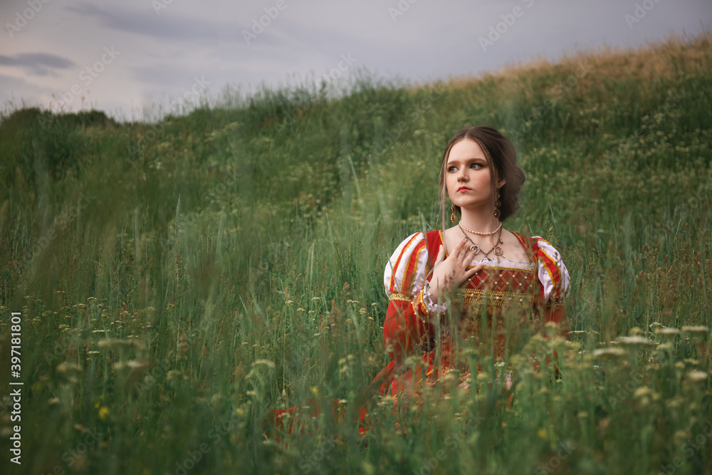 Φωτογραφία beautiful young woman in a long red medieval dress is sitting in the grass in th