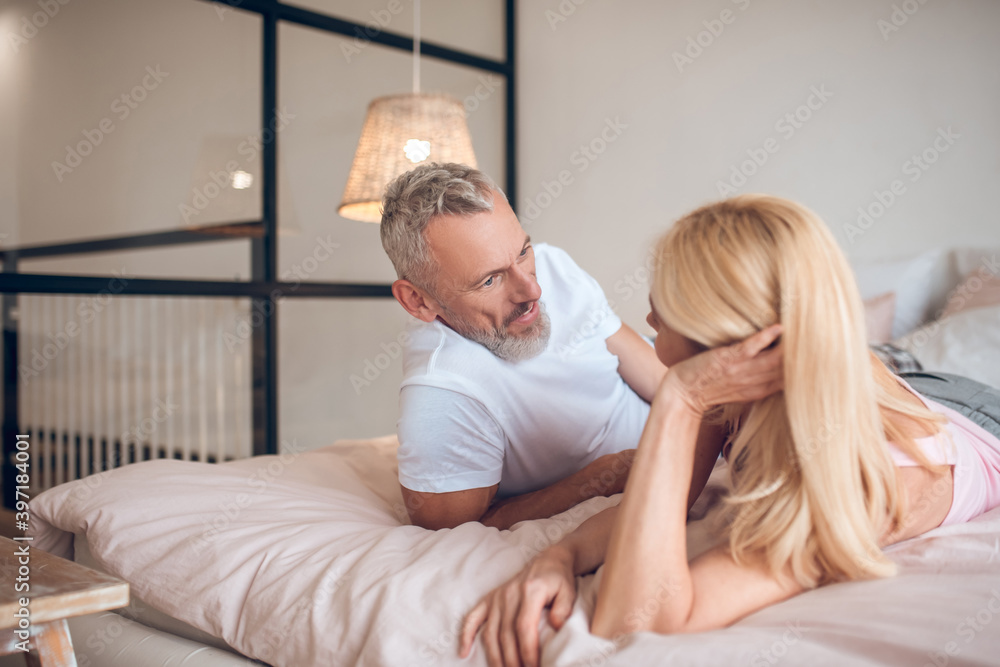Grey-haired man and a blonde woman lying on the bed and looking at each other Grey-haired man and a blonde woman lying on the bed and looking at each other