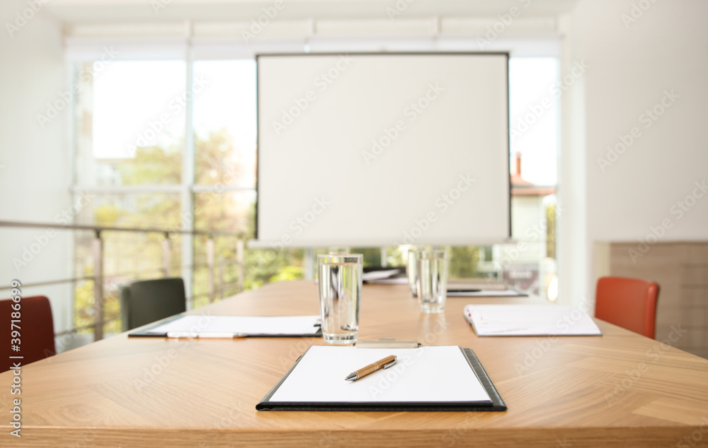 Clipboards and glasses of water on wooden table in modern office with projection screen Clipboards and glasses of water on wooden table in modern office with projection screen