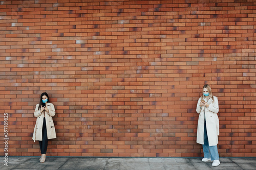 Stylish women wearing masks using smartphones while standing near brick wall in the city street. Social distance. 