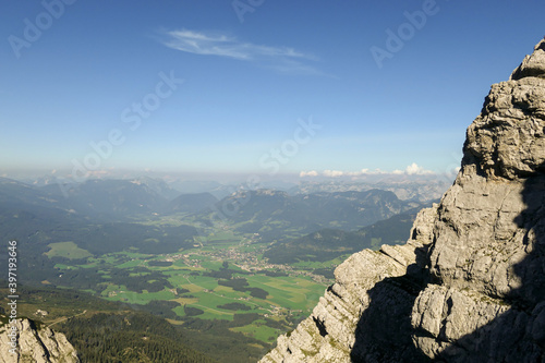 A panoramic view from an Alpine top on a vast valley. There are sharp mountains and high peaks around. The Alpine slopes are almost barren. Lush green valley. Bright day. Serenity and freedom.