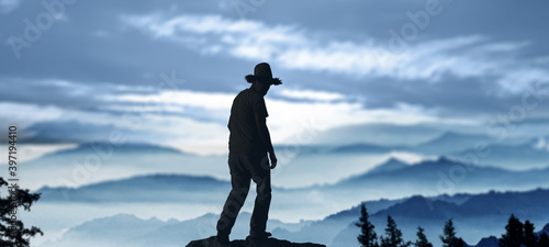 silhouette of cowboy stands on peak admiring in evening misty mountain landscape.