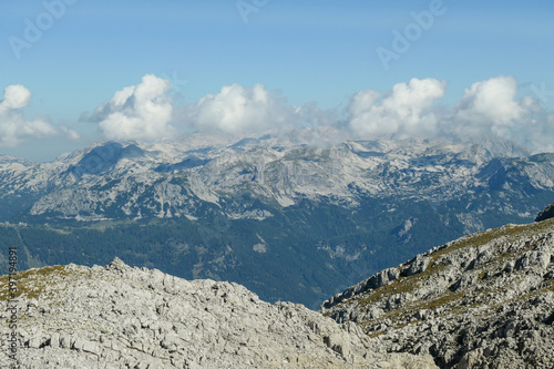A panoramic view on a distant chain of Alps from another high peak. The massive chain is partially shrouded with clouds. Below there is lush green valley and a few cities in it. Austrian landscape