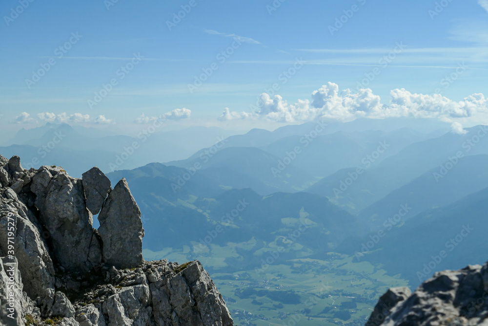 A panoramic view from an Alpine top on a vast valley. There are sharp mountains and high peaks around. The Alpine slopes are almost barren. Lush green valley. Bright day. Serenity and freedom.