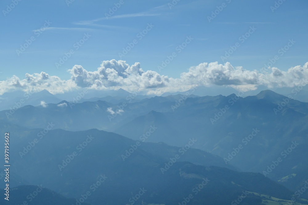 A close up view on a distant chain of Alps from another high peak. The massive chain is partially shrouded with clouds. A thin haze covering the lower parts of the mountains. Austrian landscape