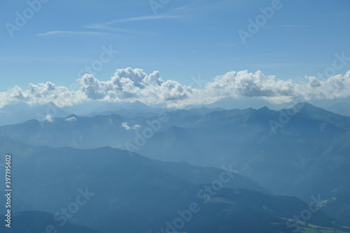 A close up view on a distant chain of Alps from another high peak. The massive chain is partially shrouded with clouds. A thin haze covering the lower parts of the mountains. Austrian landscape