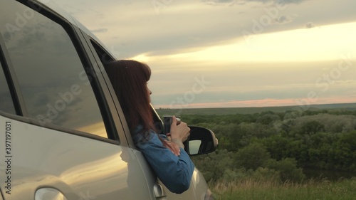 Tourist driver is resting drinking tea from mug in sun. Woman traveler holds metal mug with hot coffee in her hands and looks at sunset on street next to her car. freedom of travel and tourism by car.
