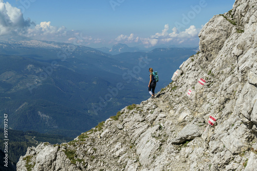 Woman walking on a very narrow and steep pathway along a mountain wall in Austrian Alp, leading to the top of Grimming. The path is marked with red-white-red sign. Dangerous trail. Following the path