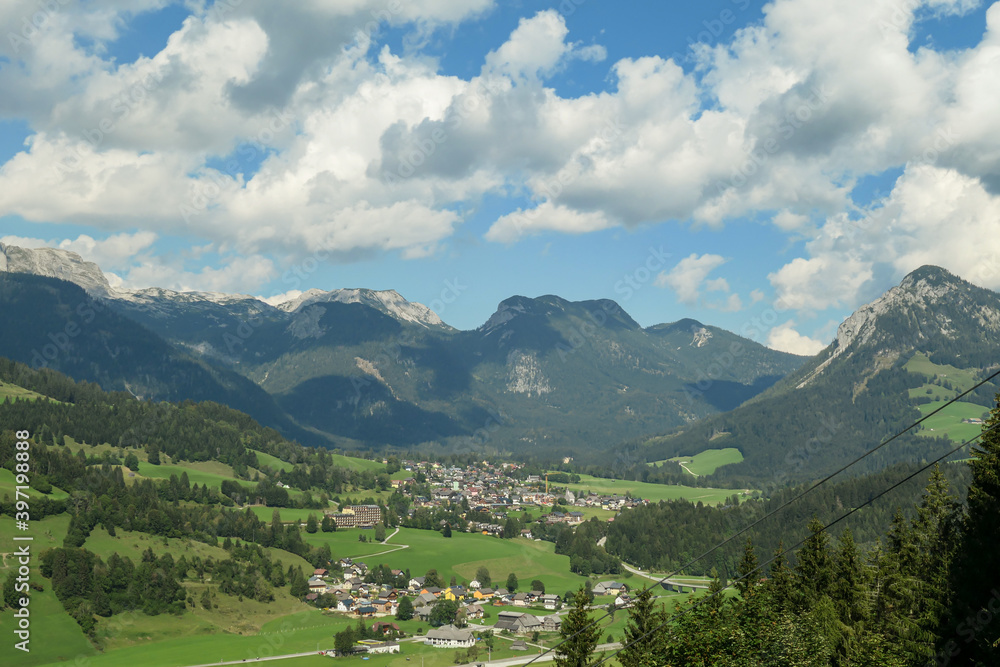 Panoramic view on a vast Alpine valley. There are sharp mountains and high peaks around. Small town at the bottom. The Alpine slopes are almost barren. Lush green valley. Bright day. Idyllic landscape
