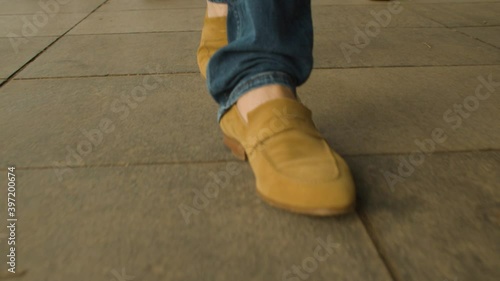 One guy walks slowly and confidently down the street in blue jeans and stylish brown sand-colored suede boots without socks. First-person view of the legs close up.