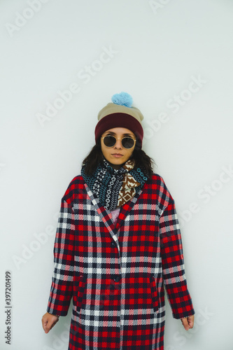 Beautiful fashion model in trendy urban street outfit. Stylish posing. Sunglasses, red coat, scarf and funny beanie.