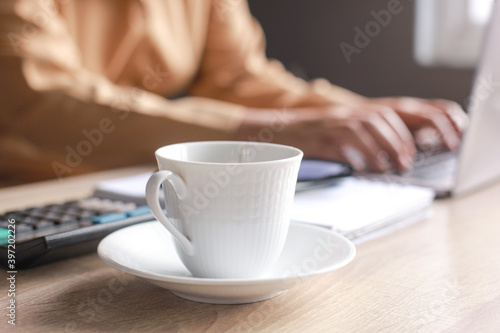 A cup of coffee on a desk office with blurry hand of businessman or worker typing on the laptop on the background