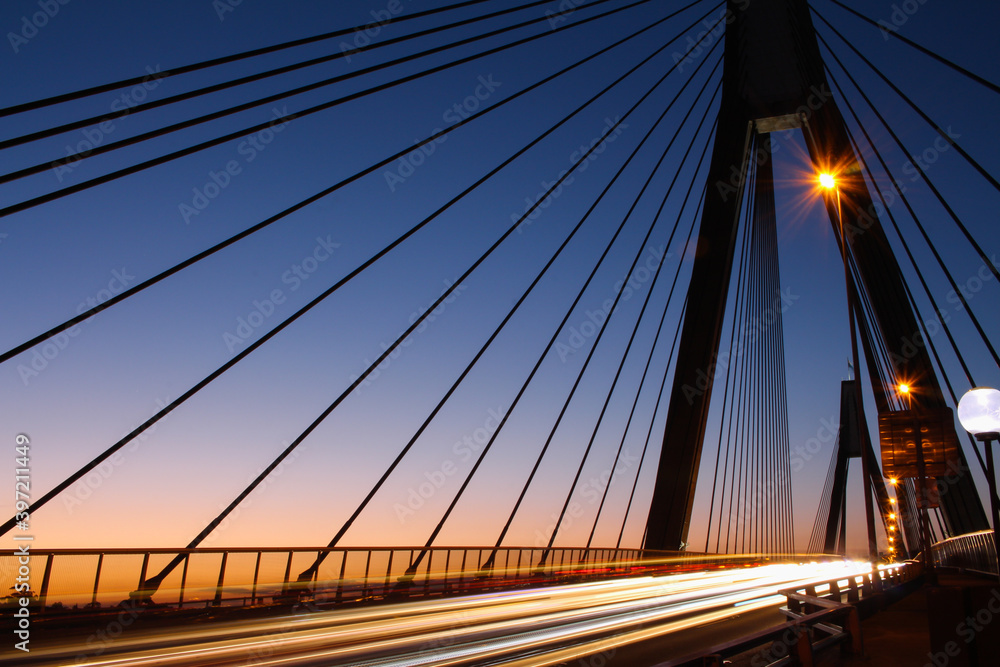 Naklejka premium Cable bridge at sunset. Long exposure, vehicle headlight trails at a cable bridge at sunset.