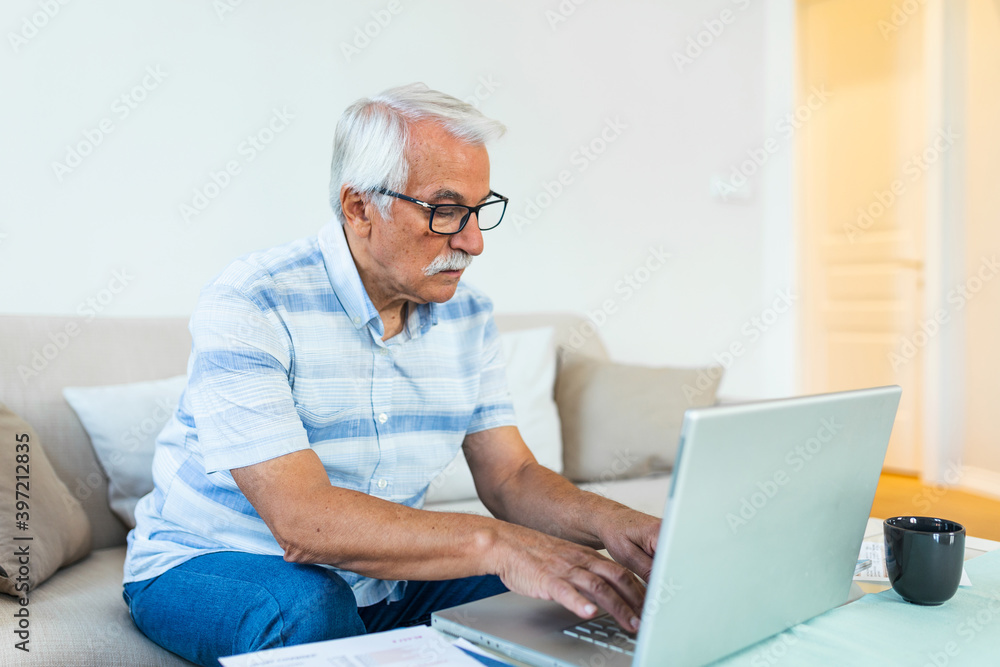 Mature businessman sit on sofa working on computer wireless device using e-banking application paying bills online, typing e-mail solve issues distantly, older generation and modern tech usage concept Mature businessman sit on sofa working on computer wireless device using e-banking application paying bills online, typing e-mail solve issues distantly, older generation and modern tech usage concept