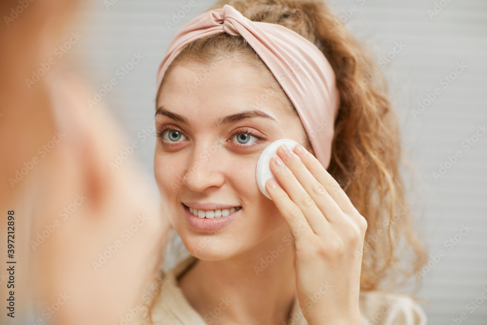 Young smiling woman looking at the mirror and removing her makeup from face with sponge Young smiling woman looking at the mirror and removing her makeup from face with sponge