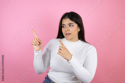 Young beautiful woman over isolated pink background surprised and pointing her fingers side © Irene