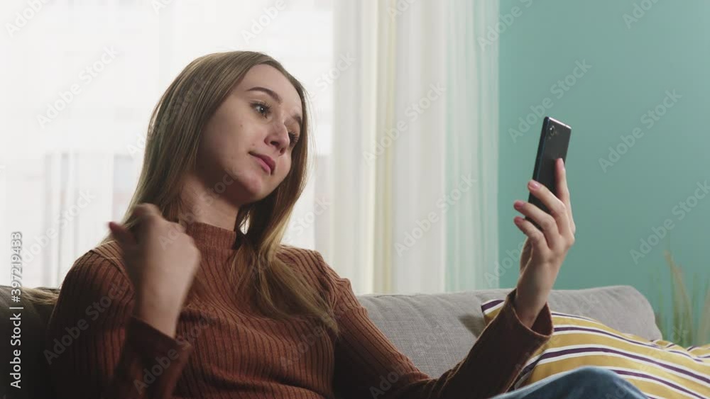 A young lady in a brown sweater and blue jeans sits on a sofa in front of a window and talks emotionally over a video call. Close-up A young lady in a brown sweater and blue jeans sits on a sofa in front of a window and talks emotionally over a video call. Close-up