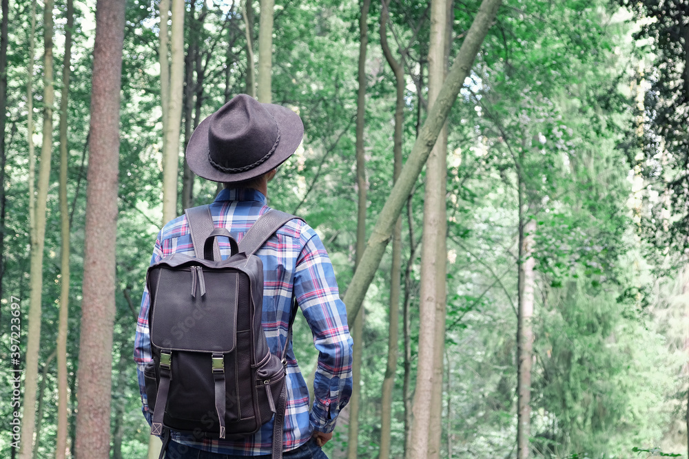Stylish young man in felt hat and plaid shirt with trendy leather backpack, standing in green forest. Escape into the wild. Adventure vibes and wanderlust. Copy space.