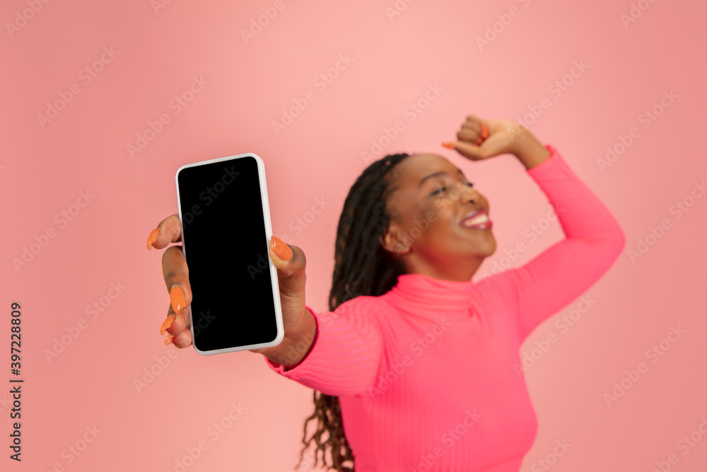 Naklejka premium Shows phone screen. Happy young beautifil african-american woman isolated on pink background. Copy space for ad, design. Half-length shot. Concept of human emotions, facial expression.